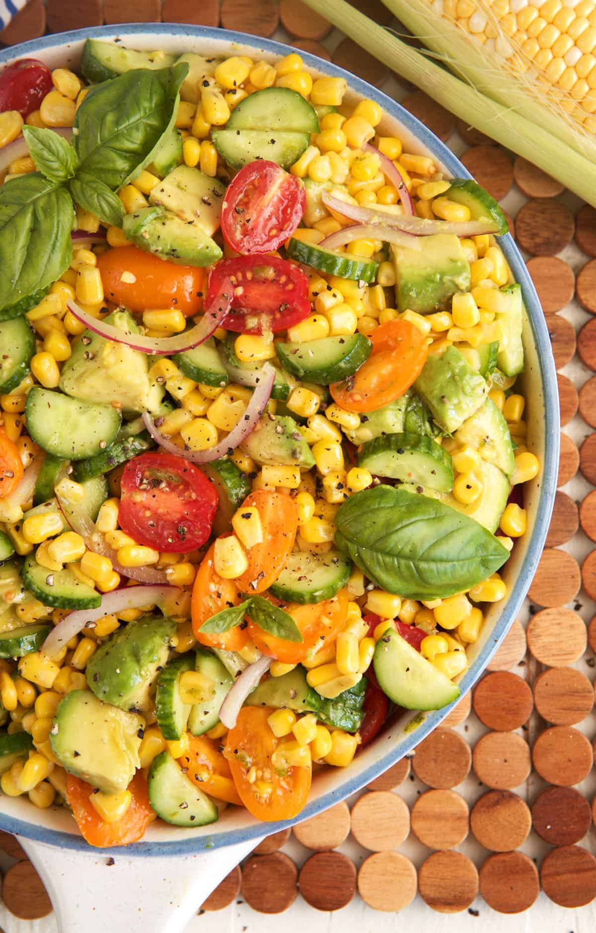 Overhead view of avocado corn salad in bowl with tomatoes, cucumbers, and fresh basil