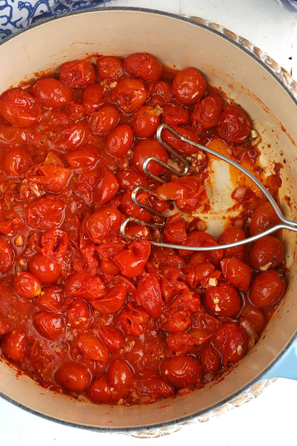 Overhead view of roasted tomato sauce in pot, with potato masher crushing tomatoes