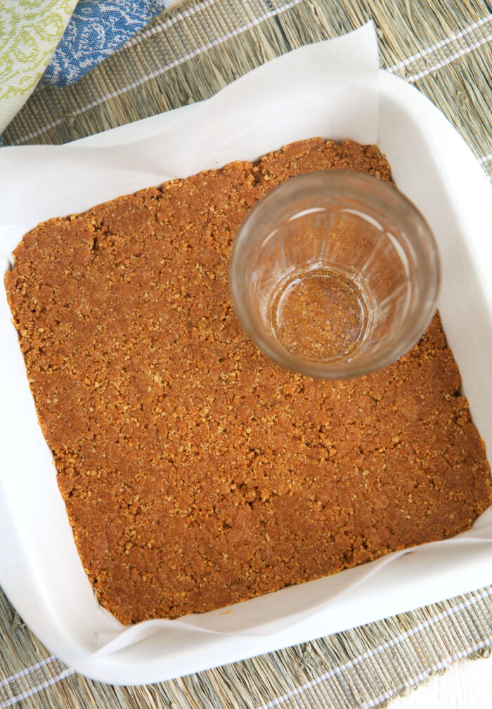 graham cracker crust in a white baking dish with a glass on top for pressing the crust into the dish.
