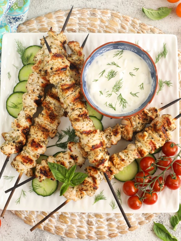 Overhead view of chicken souvlaki on platter with tzatziki, cucumbers, and cherry tomatoes