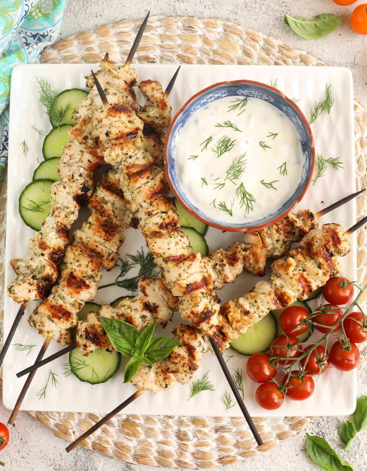 Overhead view of chicken souvlaki on platter with tzatziki, cucumbers, and cherry tomatoes