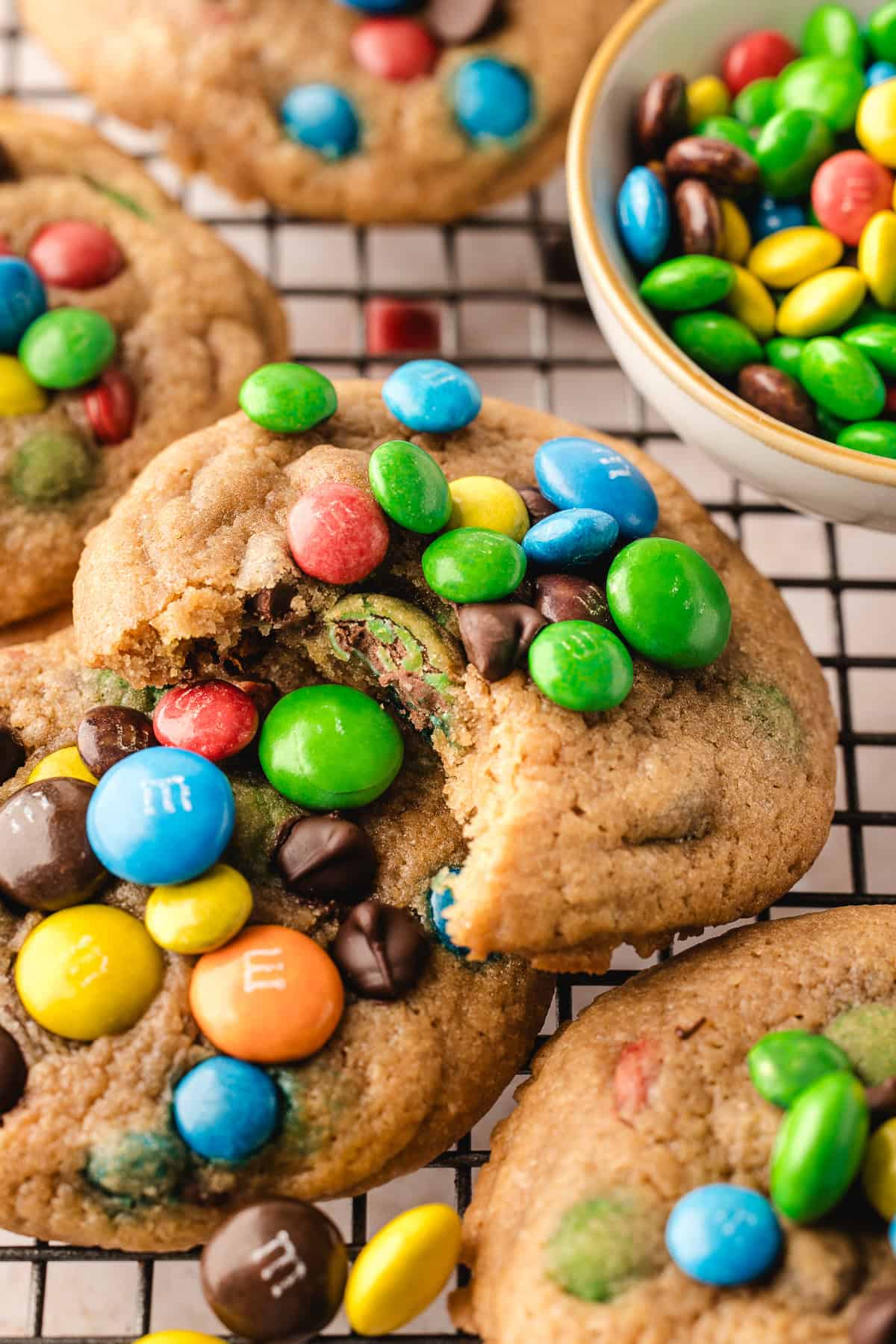 Soft and chewy chocolate chip M&M cookies on wire rack, with bite taken out of one to show texture