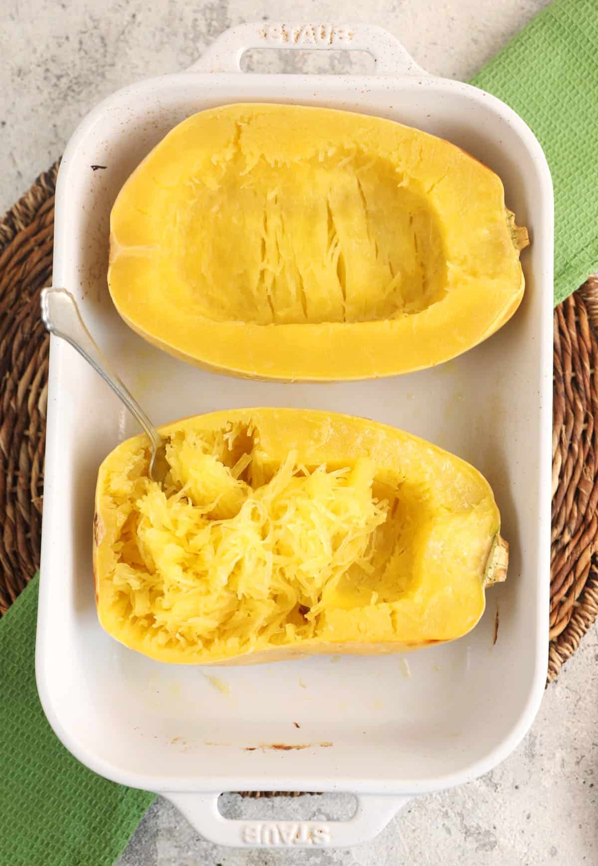 Halved roasted spaghetti squash in baking dish with fork shredding on half