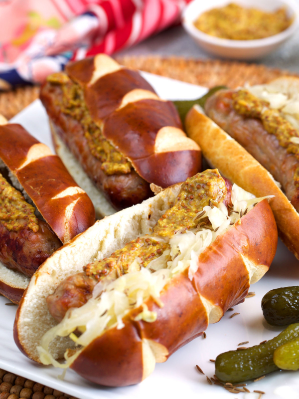 Air fryer brats on pretzel rolls with sauerkraut and mustard on a white platter.