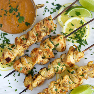 Grilled Chicken Satay on a white square platter with a bowl of peanut sauce and sliced limes.