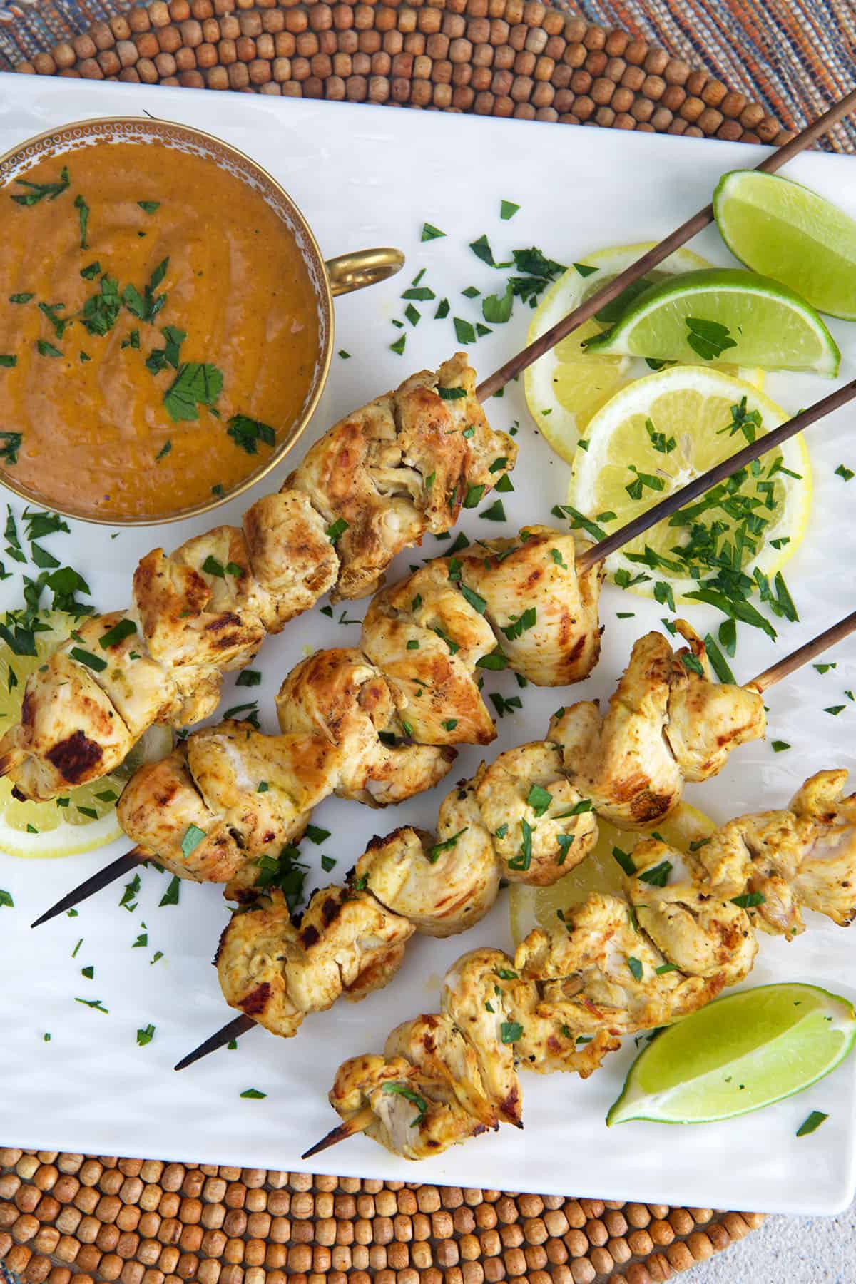 Grilled Chicken Satay on a white square platter with a bowl of peanut sauce and sliced limes.