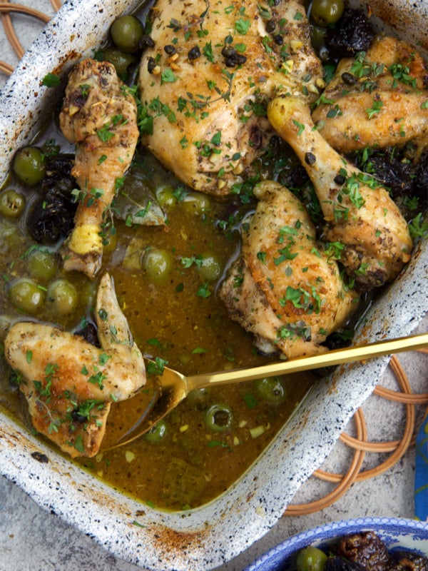 Chicken Marbella in a baking dish with a gold serving spoon.