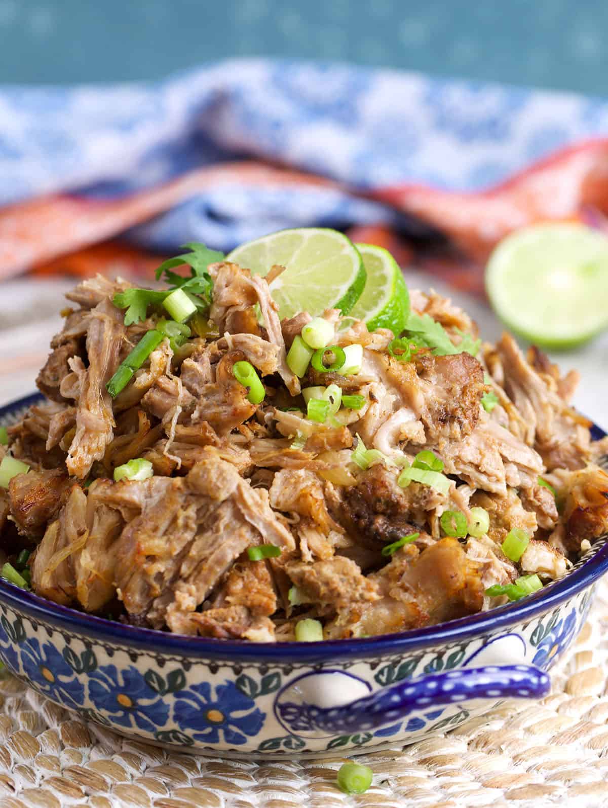 bowl of crispy pork carnitas on a wicker place mat topped with lime slices.