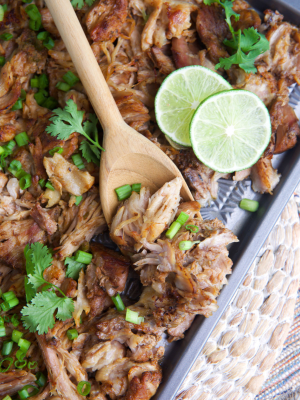 Pork carnitas on a baking sheet with limes, cilantro and green onion sprinkled on top and a serving spoon.