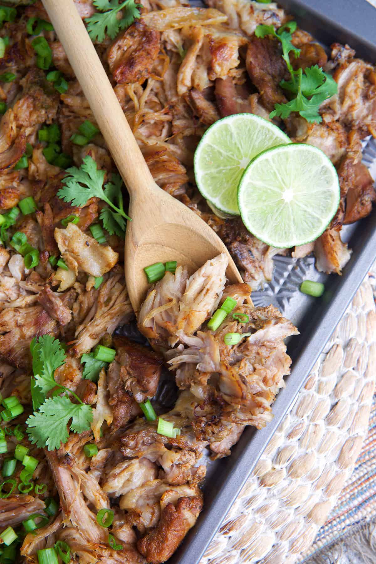 Pork carnitas on a baking sheet with limes, cilantro and green onion sprinkled on top and a serving spoon.