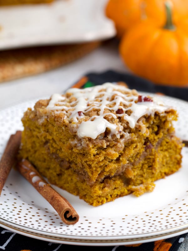 Pumpkin crumb Cake slice on a white plate with cinnamon sticks and a couple mini pumpkins in the background