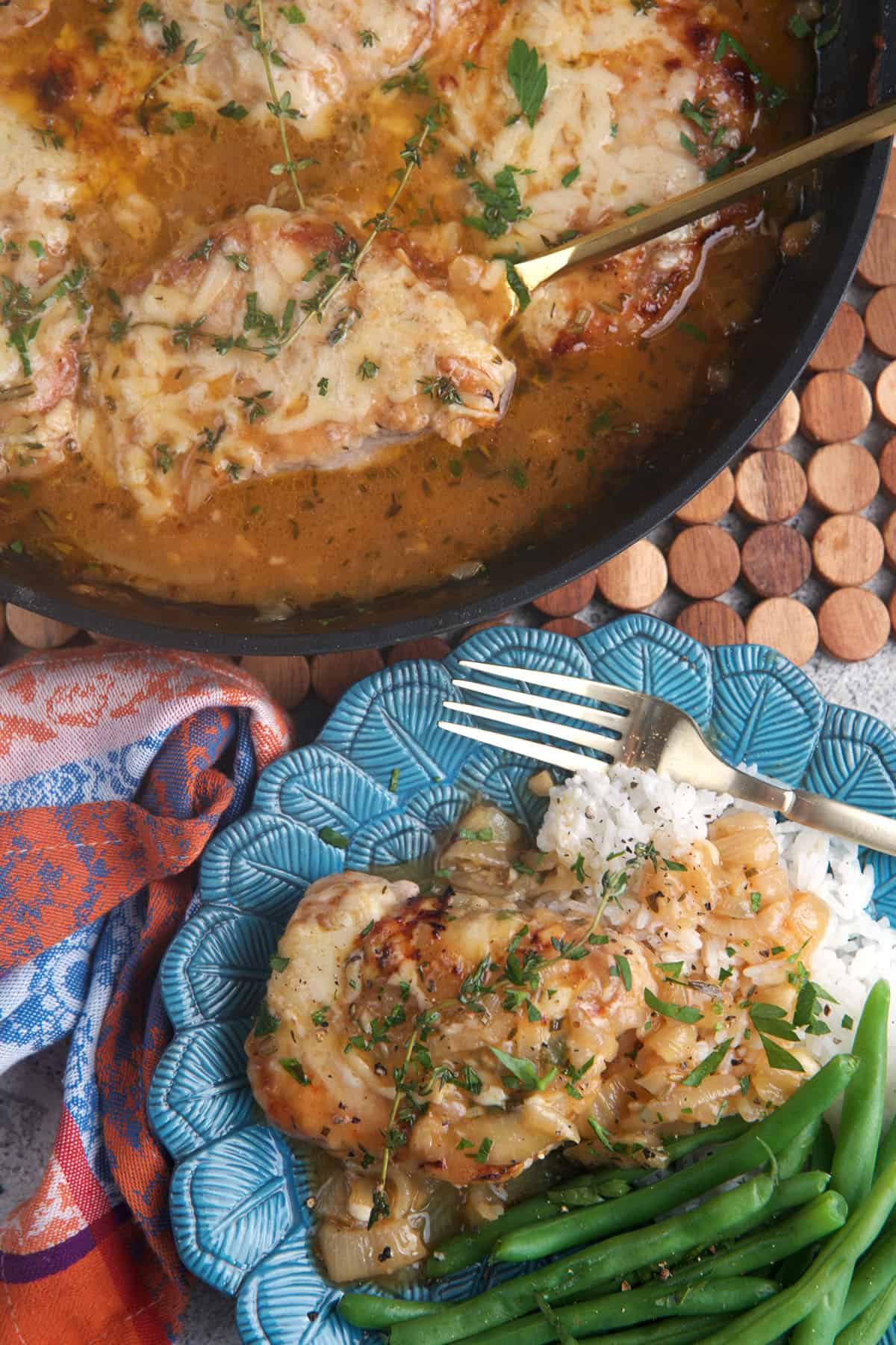 French onion pork chops in a skillet with one chop on a blue plate with green beans