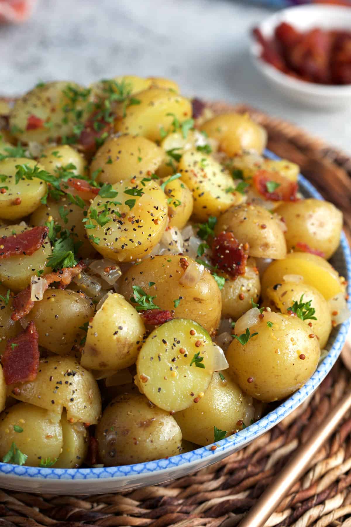 German Potato Salad recipe in a round serving bowl with a small dish or bacon in the background.