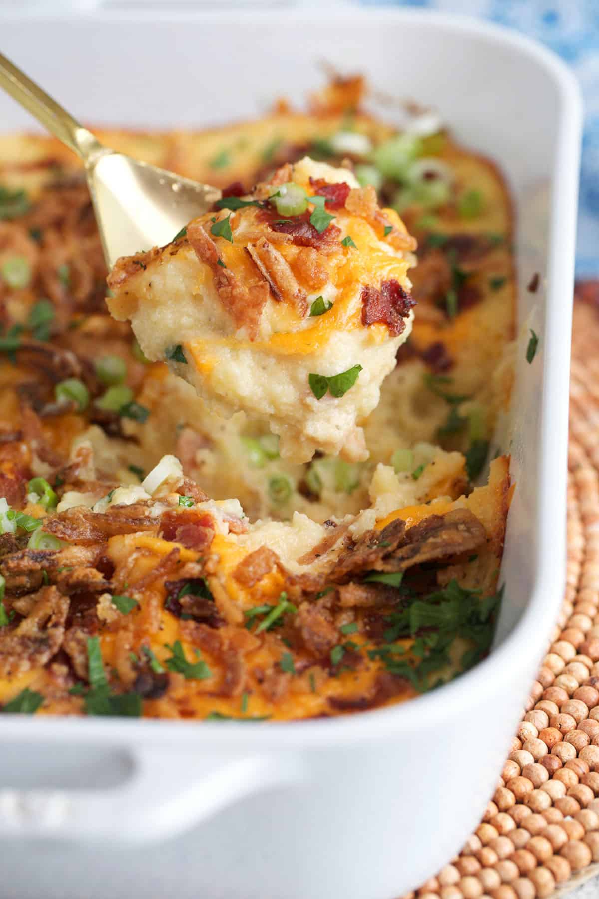Loaded Mashed Potato Casserole in a white staub casserole dish with a gold serving spoon serving a portion.
