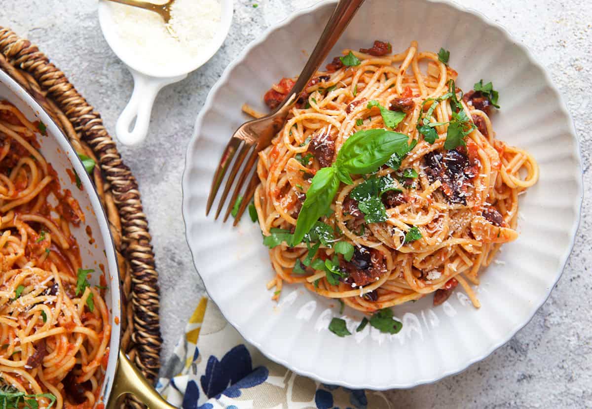 spaghetti puttanesca in a white bowl with a dish or parmesan cheese