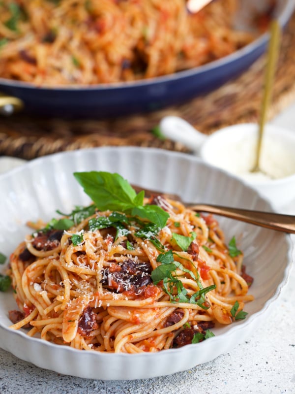 Pasta puttanesca in a white bowl with a skillet of pasta in the background