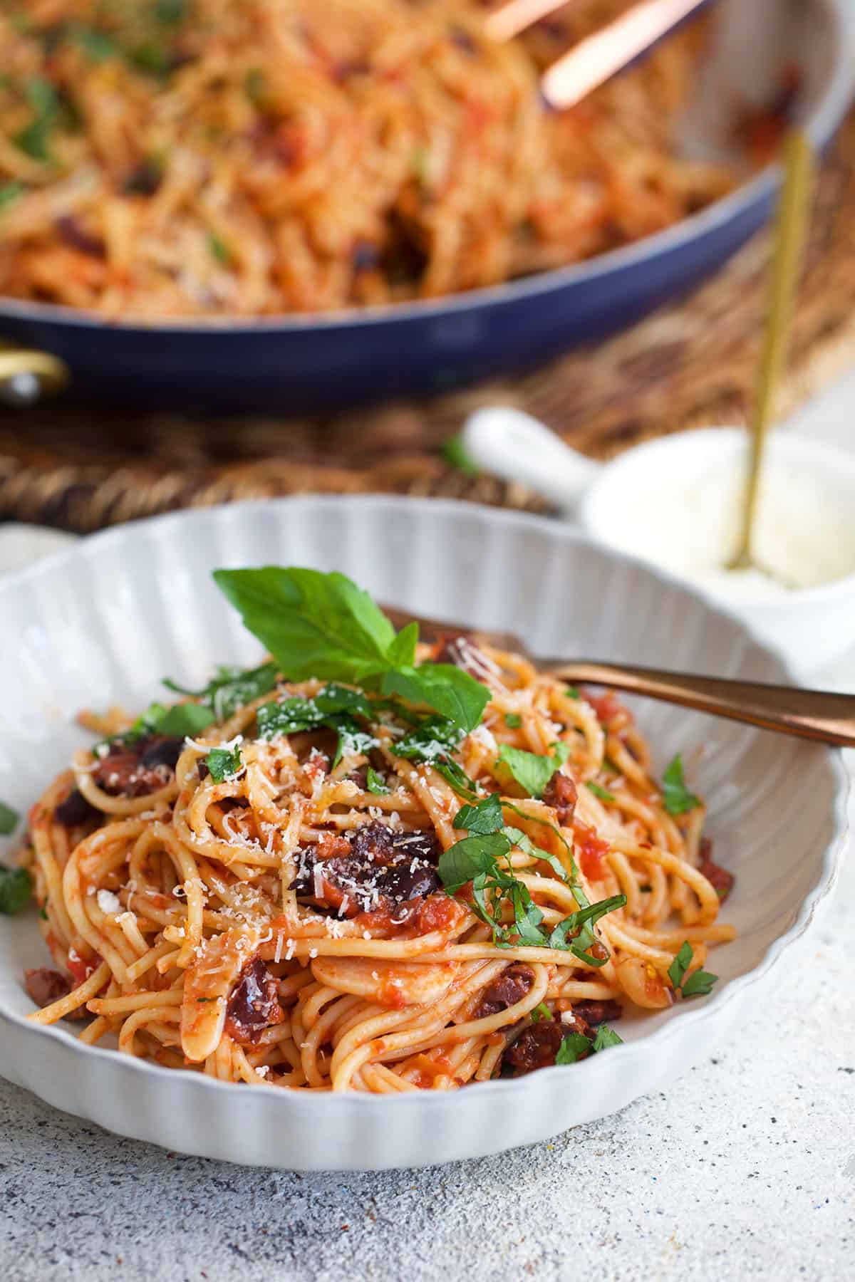 Pasta puttanesca in a white bowl with a skillet of pasta in the background