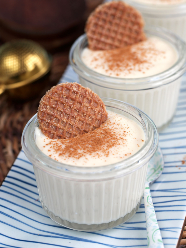 Vanilla pudding in glass jars on a blue and white striped tray. Each is garnished with cinnamon and a mini stroopwafel.