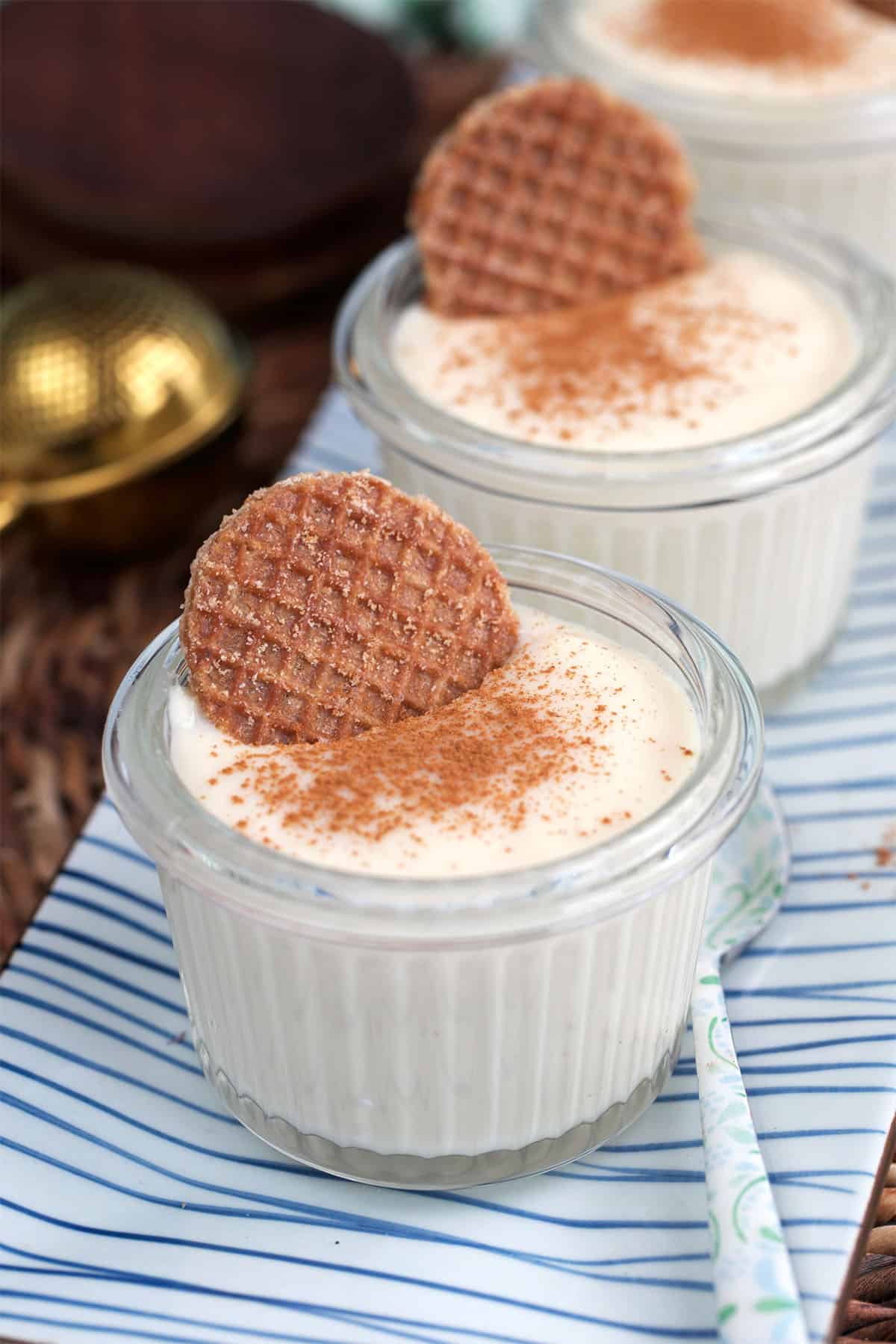 Vanilla pudding in glass jars on a blue and white striped tray. Each is garnished with cinnamon and a mini stroopwafel.