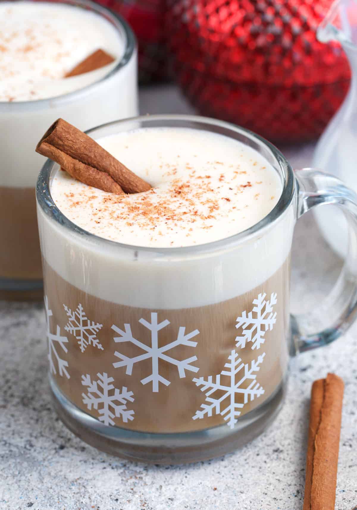 Eggnog Latte in a glass mug with snowflakes on it and a cinnamon stick on top.
