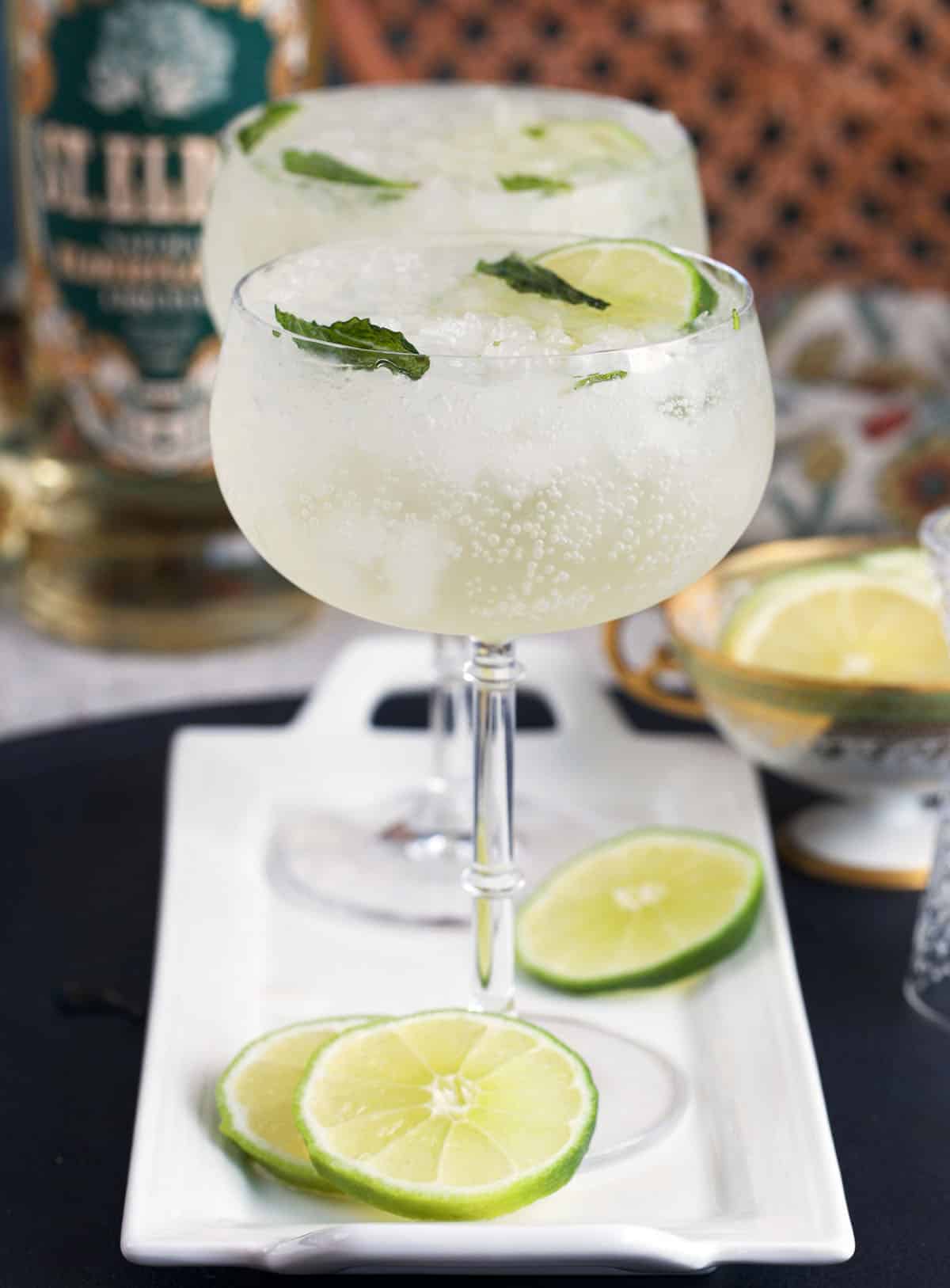 Two coupe glasses with elderflower spritz on a white tray with limes and mint leaves.