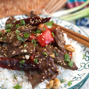 Kung Pao Beef on a bed of rice plated on a white and turquoise plate with wooden chopsticks.