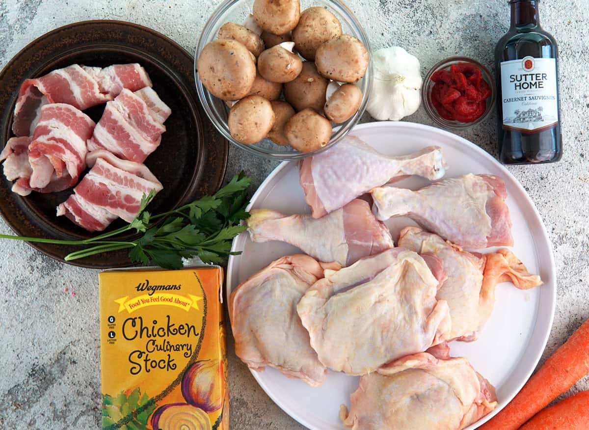 Ingredients for coq au vin, bacon, mushrooms, chicken stock, chicken parts, wine, onion, carrots and garlic.