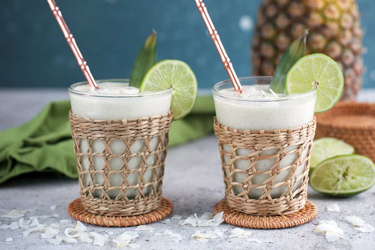 Two rattan glasses filled with pineapple smoothie with a pineapple in the background.