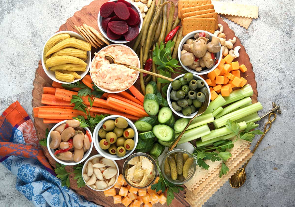 Classic Relish Tray shown on a wood round board with assorted dips in white bowls, vegetable spears, nuts and pickled items served with herbs and olives.