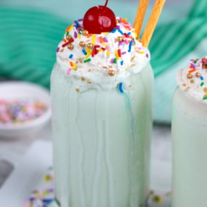 Shamrock Shake in a glass with whipped cream, sprinkles and a cherry. Two gold straws are inside the shake.