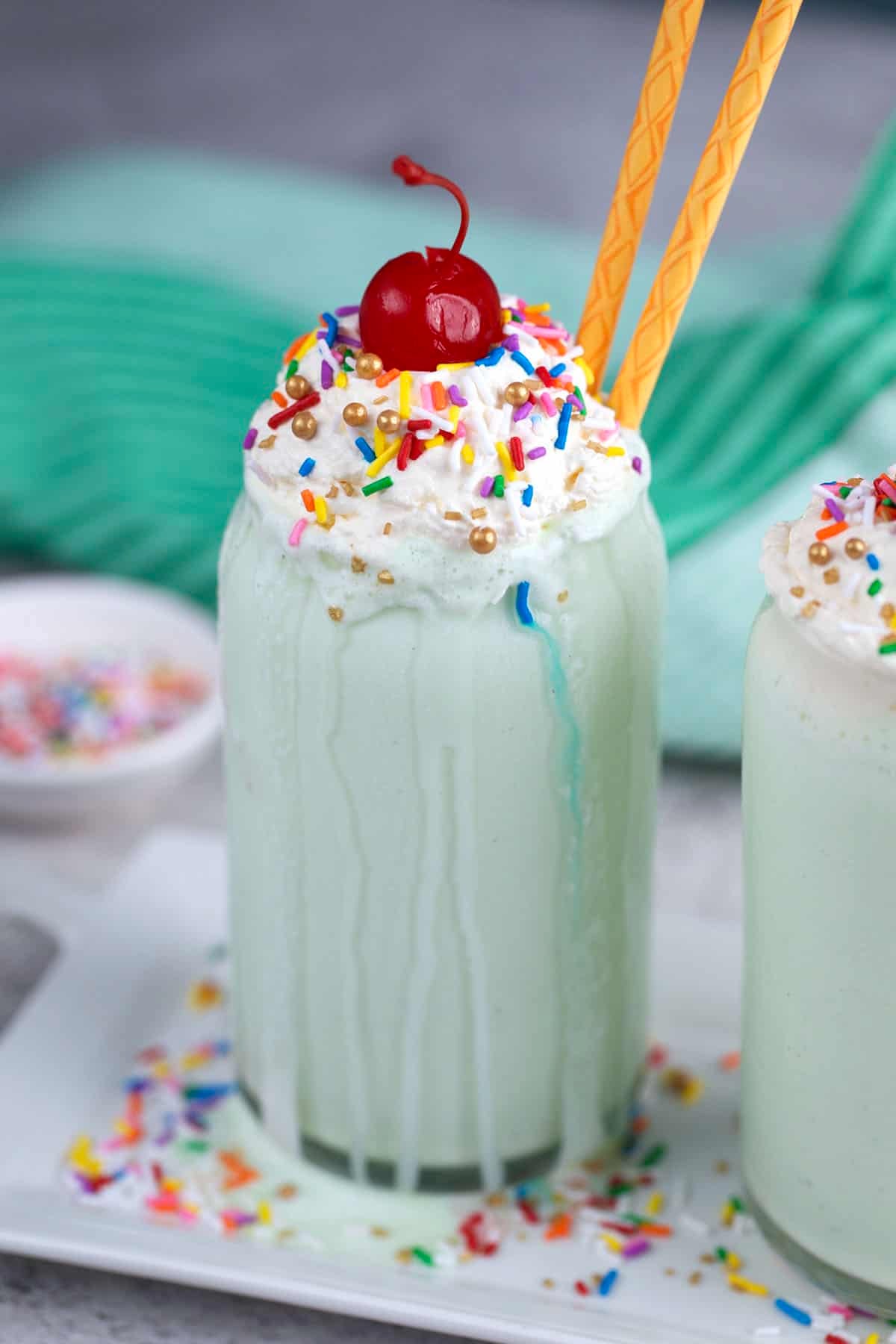 Shamrock Shake in a glass with whipped cream, sprinkles and a cherry. Two gold straws are inside the shake.