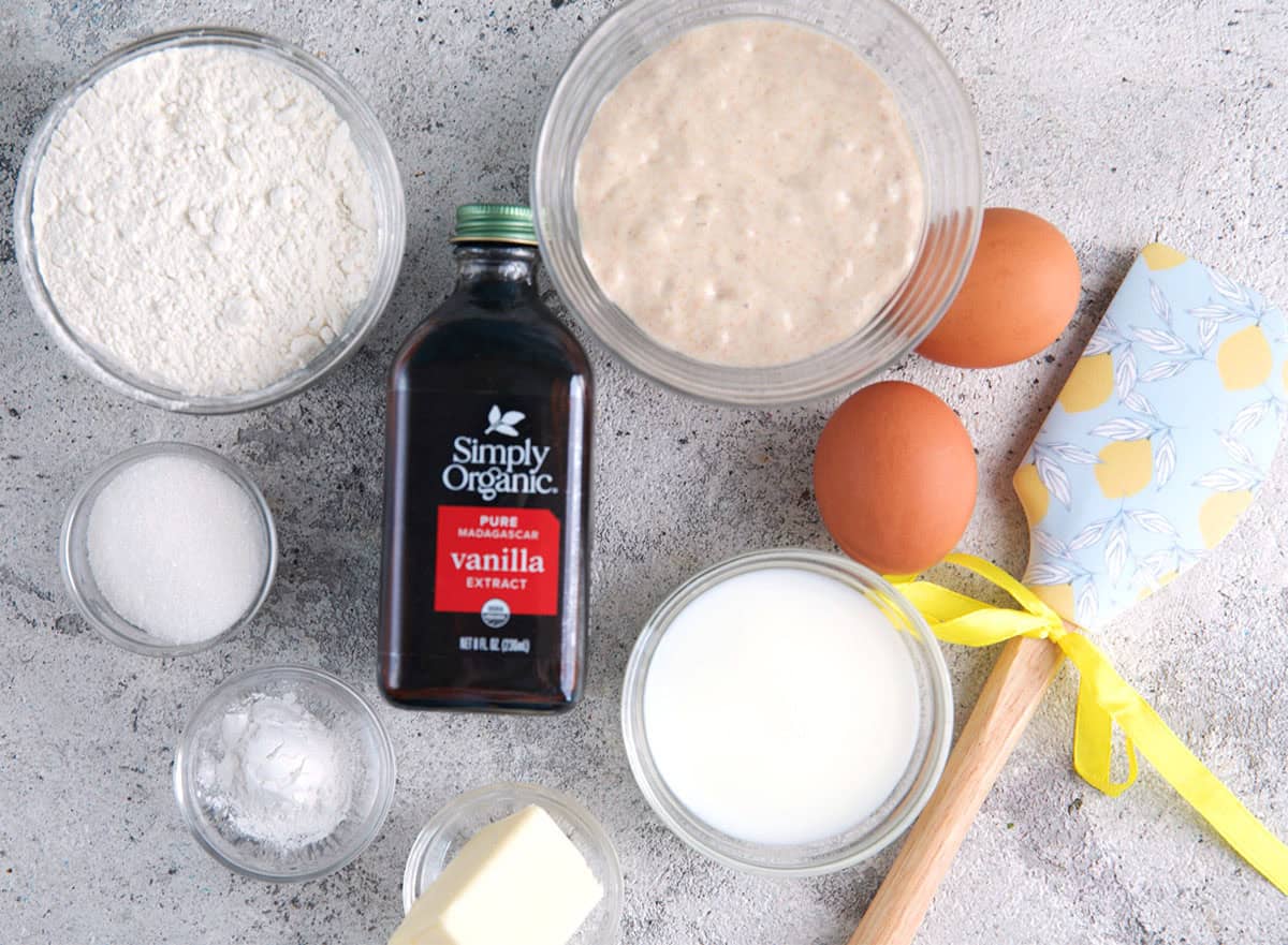 Ingredients for sourdough pancakes including flour, baking powder, baking soda, sugar, butter, vanilla, eggs and sourdough starter discard with a spatula.