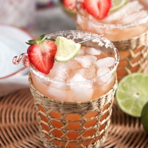 Strawberry Margarita on the rocks in a glass with a rattan holder and topped with a strawberry and lime cocktail skewer