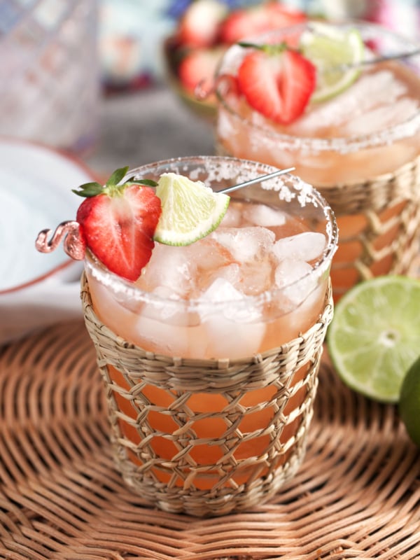Strawberry Margarita on the rocks in a glass with a rattan holder and topped with a strawberry and lime cocktail skewer