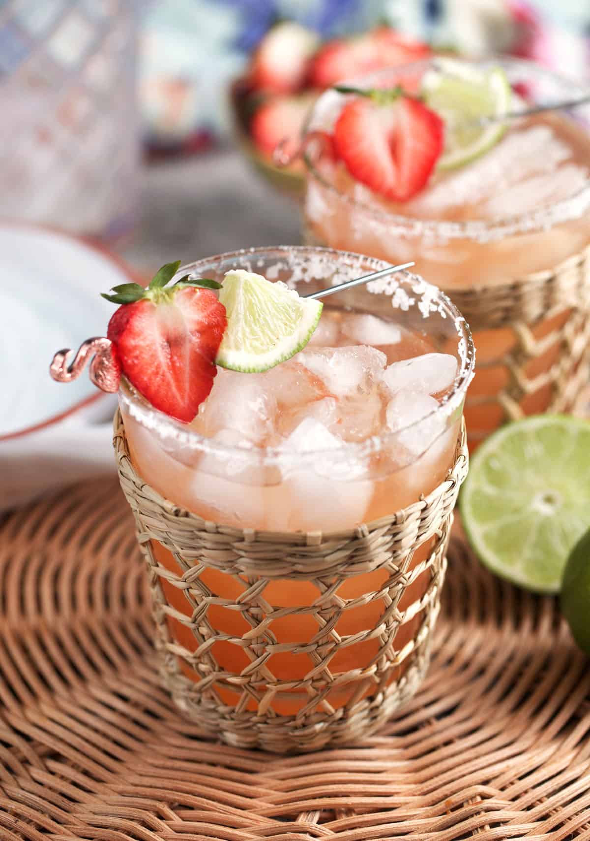 Strawberry Margarita on the rocks in a glass with a rattan holder and topped with a strawberry and lime cocktail skewer