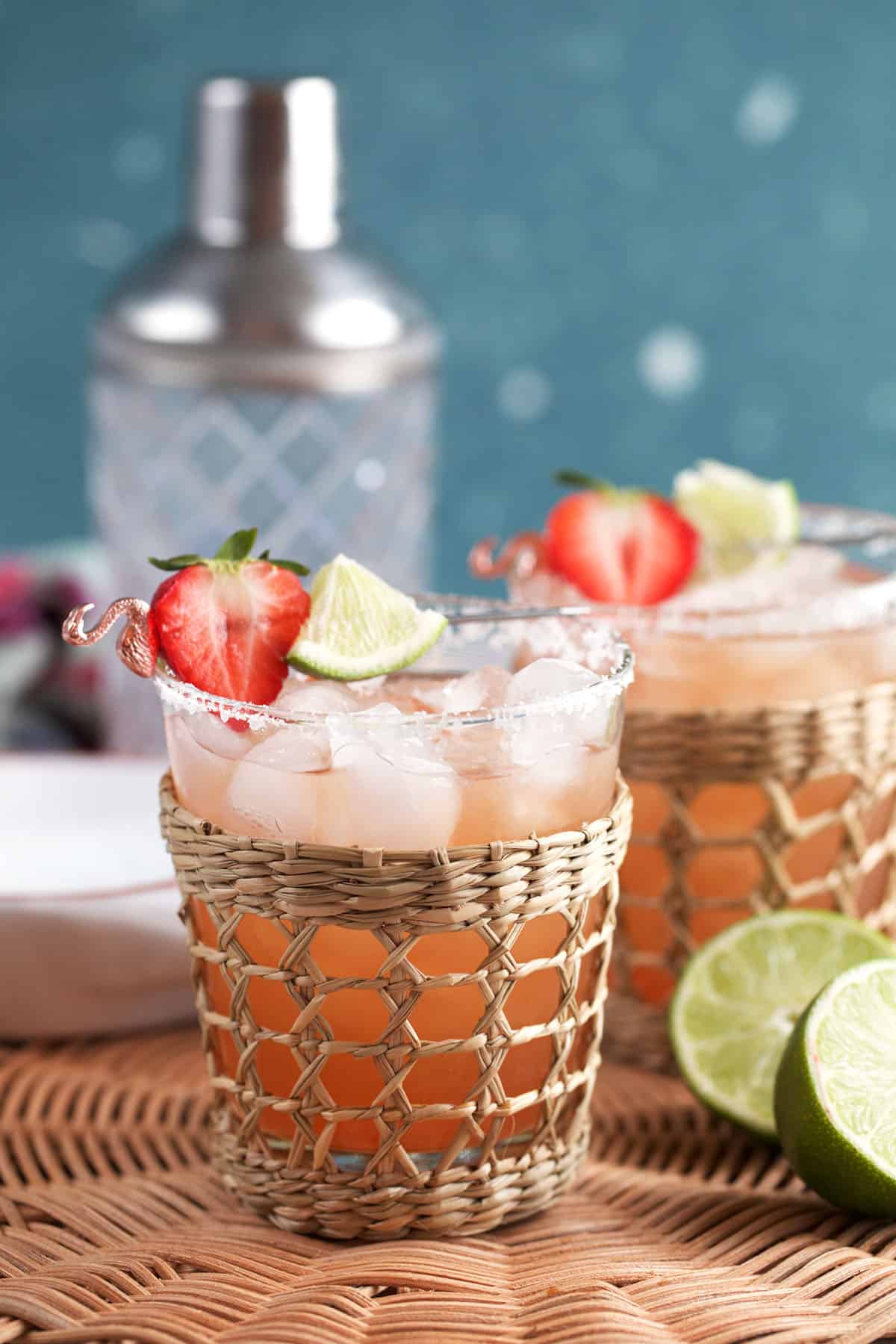 two strawberry margaritas in rattan glasses with a cocktail shaker in the background.