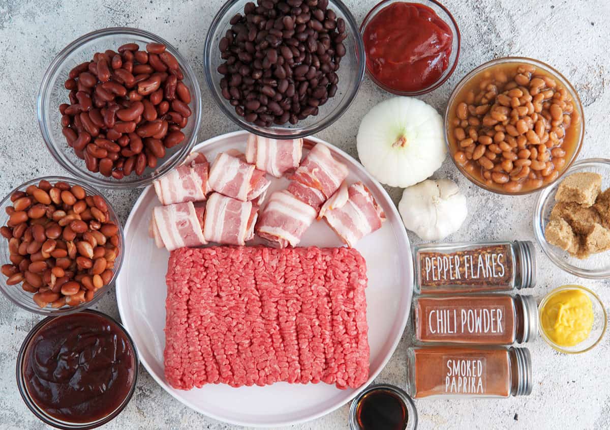 Ingredients for Cowboy Beans on a gray background. beans in bowls with ground beef and bacon on a plate and spices arranged on the sides.