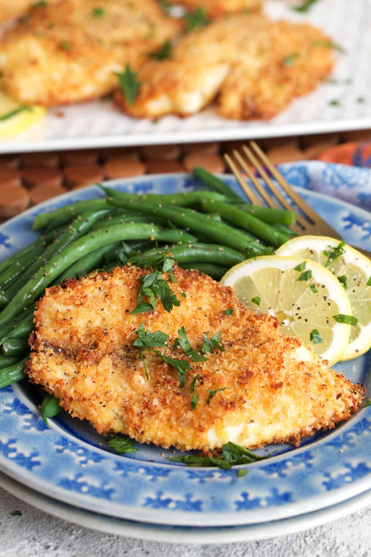 parmesan crusted tilapia with green beans and a lemon on a blue plate.