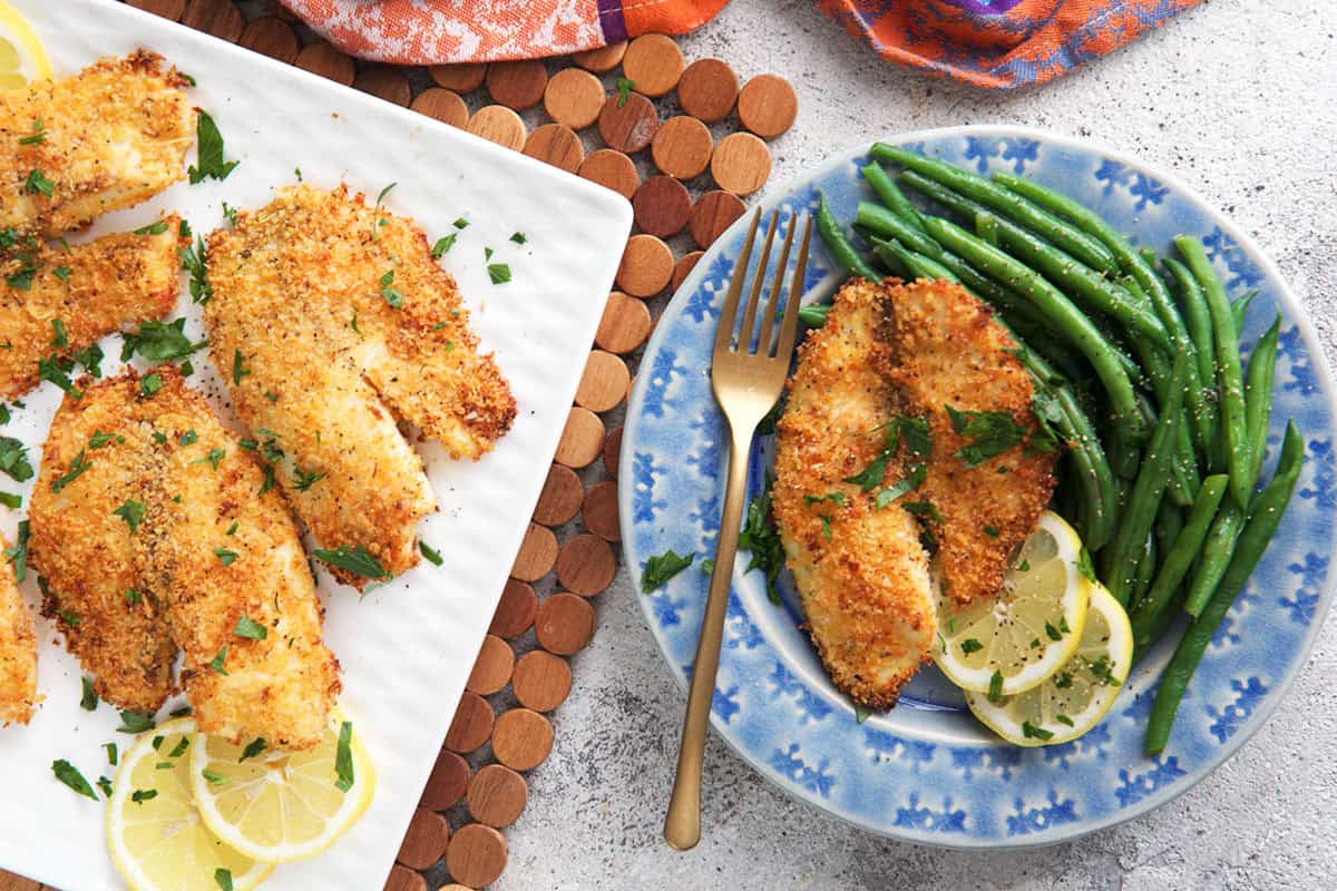 Parmesan Crusted Tilapia on a plate with green beans beside a platter of baked tilapia.