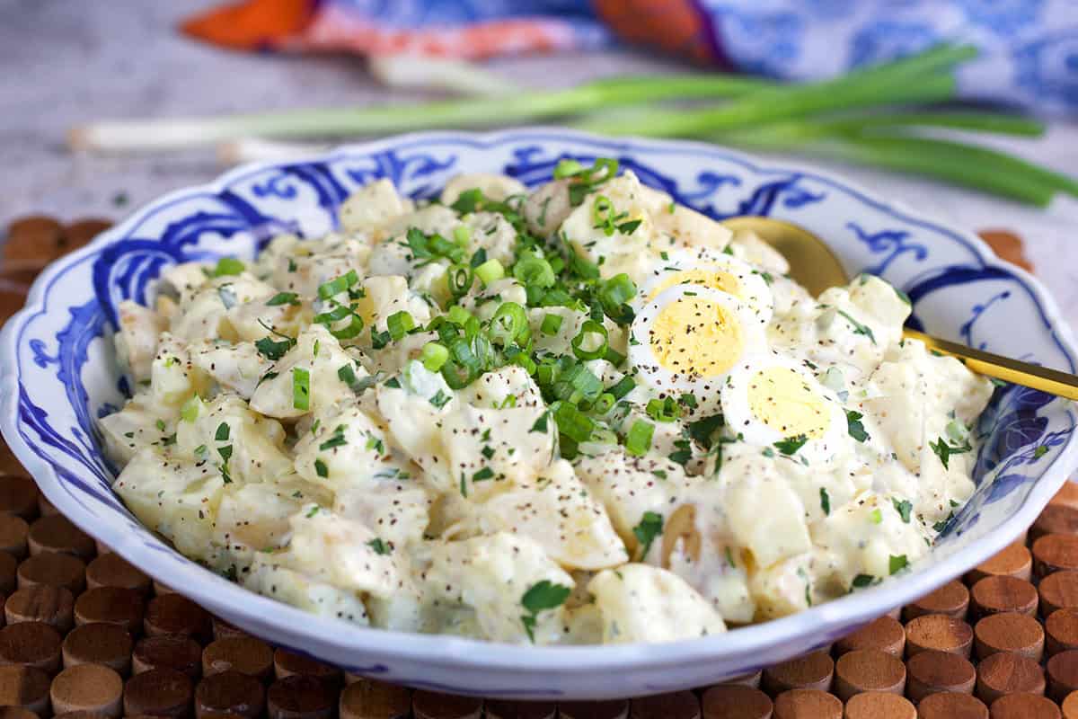 Amish potato salad recipe topped with sliced eggs and chopped herbs in a blue and white serving bowl