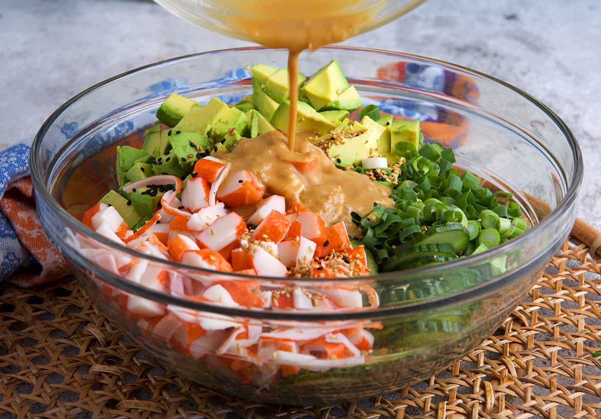 dressing being poured over ingredients for California roll cucumber salad in a glass mixing bowl