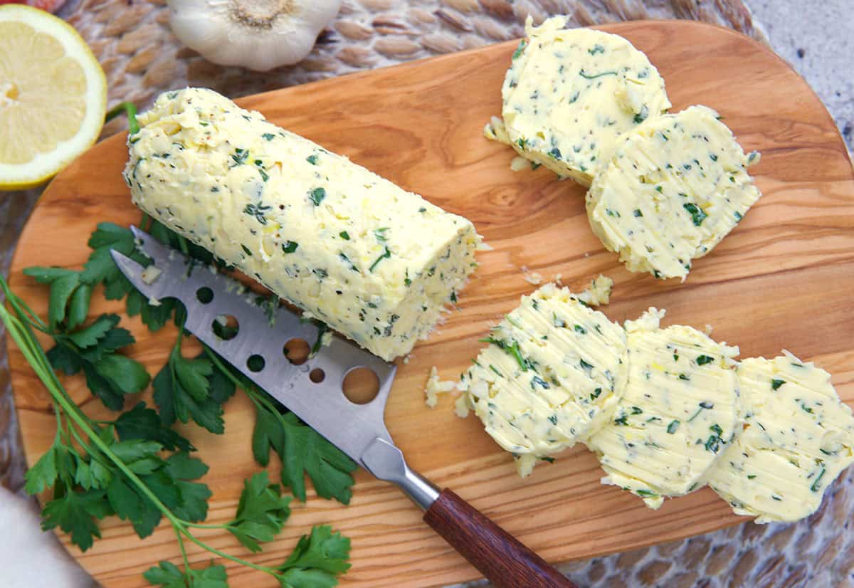 Cowboy Butter on an olive wood board cut into rounds with fresh herbs and a lemon