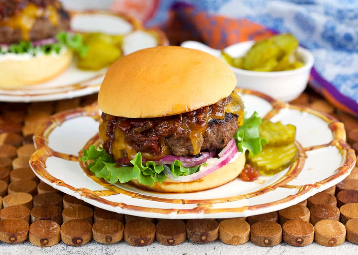 Crack Burger on a soft burger bun topped with bacon jam plated on a white plate with a side of bread and butter pickles.