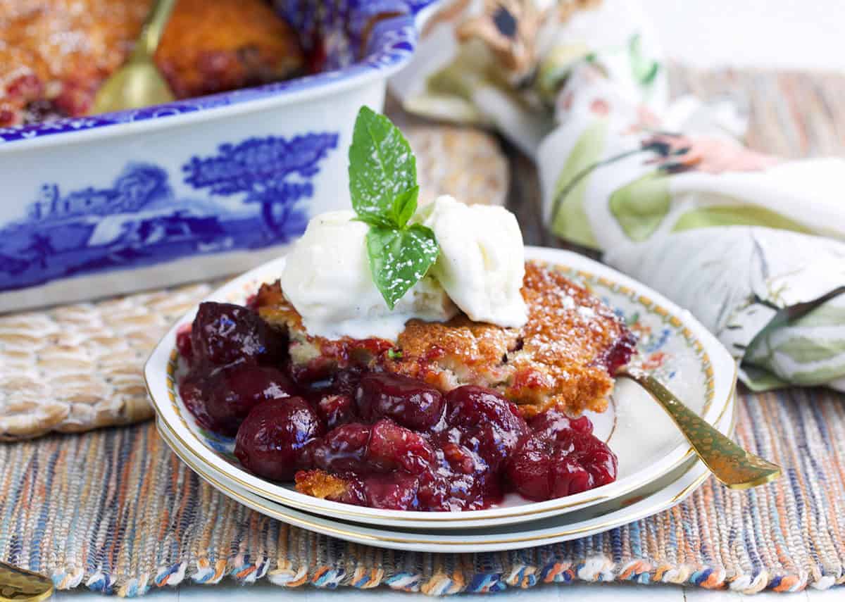 Cherry cobbler topped with vanilla ice cream with a baking dish in the background.