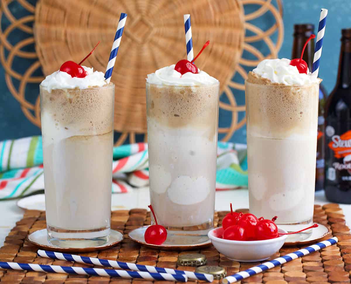 Three glasses of root beer float with striped straws and a bowl of cherries.