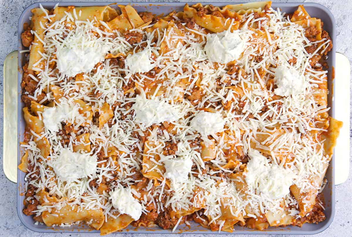 Broken lasagna noodles tossed with meat sauce on a sheet pan topped with cheese ready to be baked in the oven