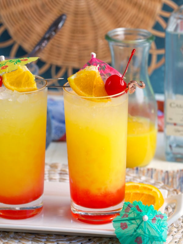 Tequila Sunrise cocktail in a collins glass with an orange slice, cherry and drink umbrella on top