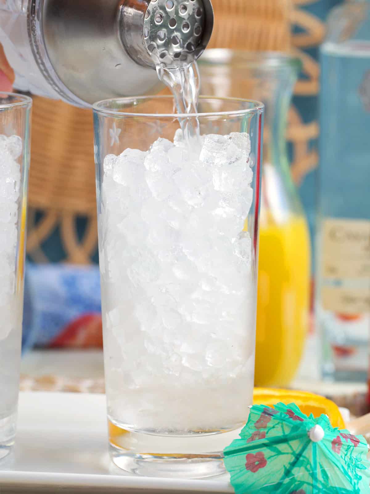 collins glass filled with pellet ice and tequila being poured into the glass with a cocktail shaker