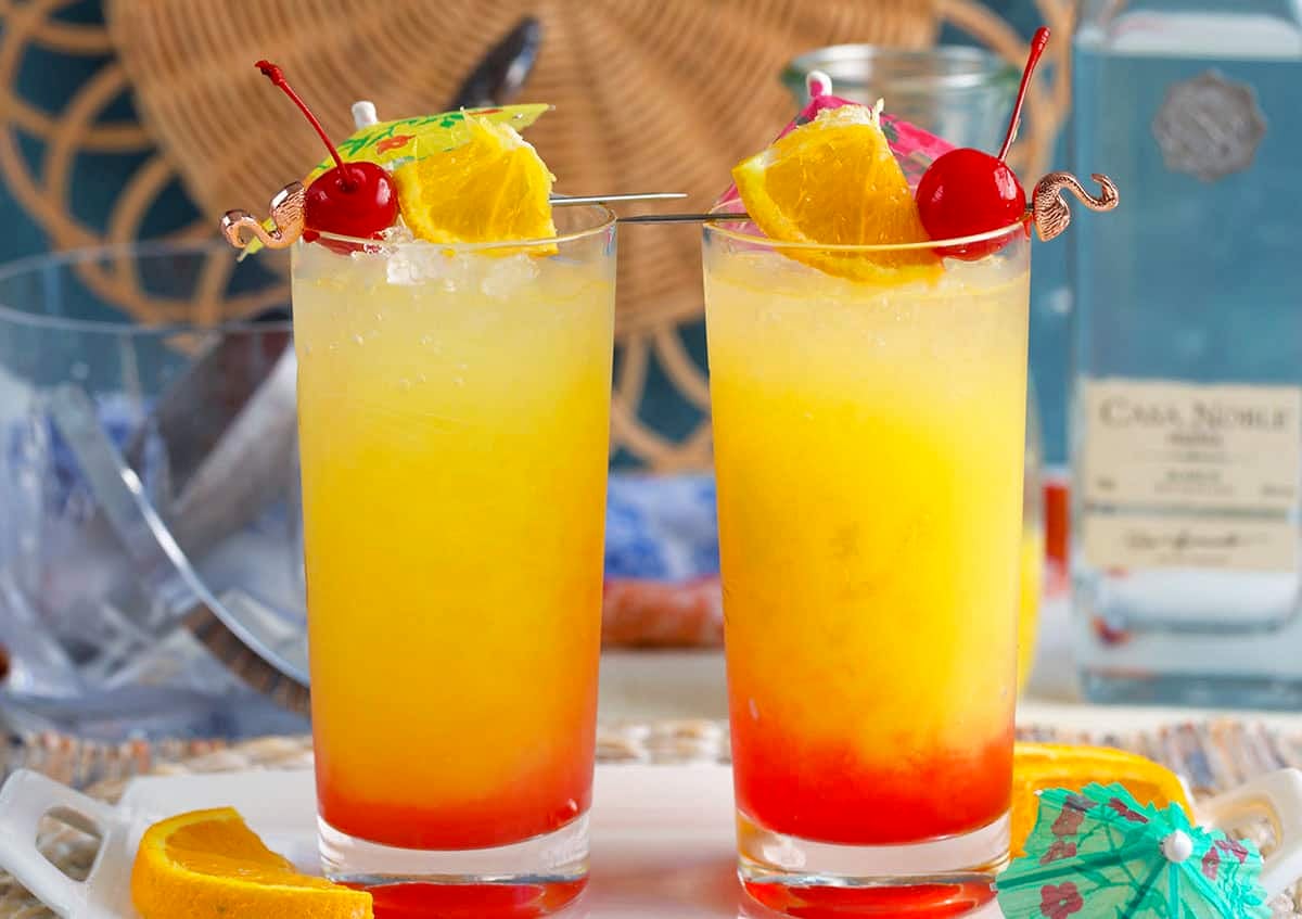 two glasses of tequila sunrise cocktails with tropical garnishes.