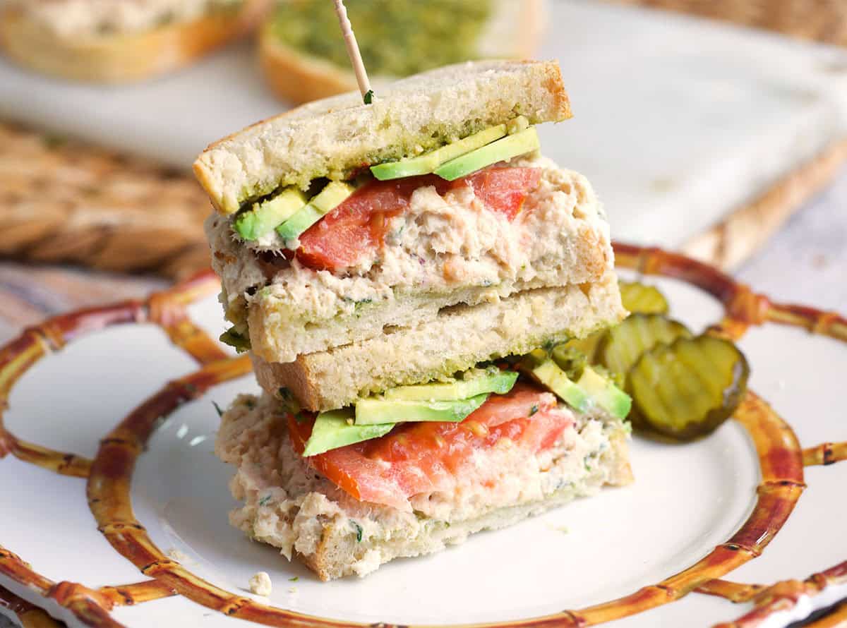 Two halves of a tunacado sandwich stacked on a white plate with pickles.
